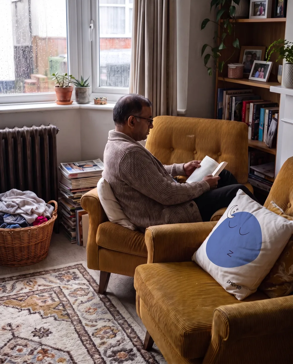 A man reads in a mustard armchair on a rainy day, with the Émomio Tiredness cover tucked behind his back.